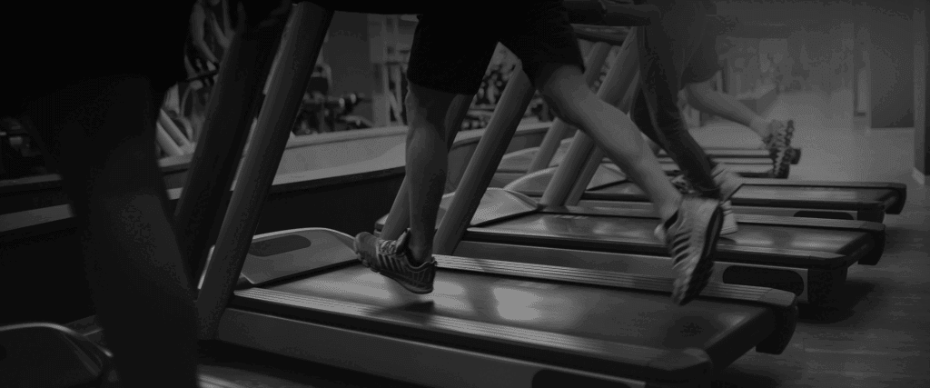 Gros plan en noir et blanc sur les jambes et chaussures de plusieurs personnes courant intensément sur des tapis de course dans une salle de sport.