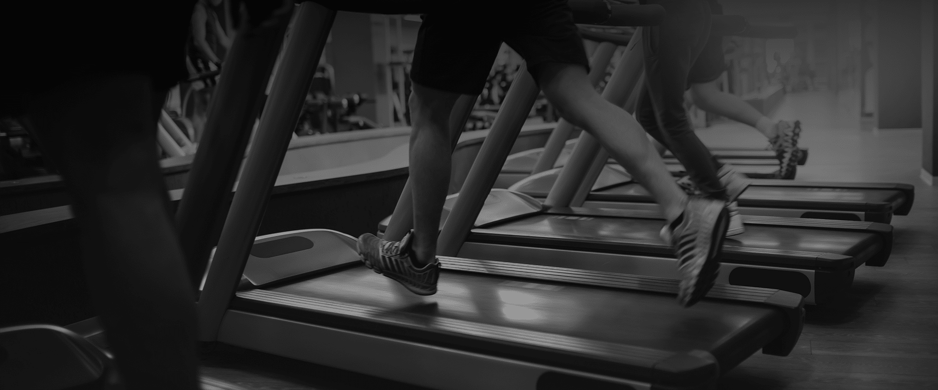 Gros plan en noir et blanc sur les jambes et chaussures de plusieurs personnes courant intensément sur des tapis de course dans une salle de sport.