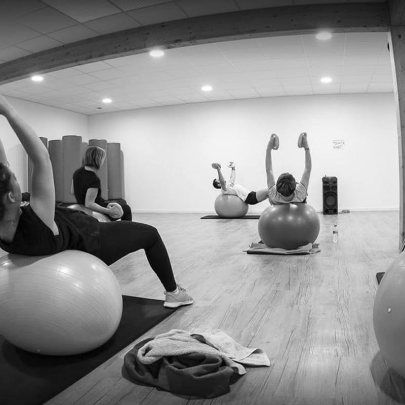 Entraînement avec ballons de stabilité : cours de fitness Exercices de gainage avec ballons de stabilité (Swiss balls) en noir et blanc, quatre personnes s'entraînent dans une salle lumineuse.