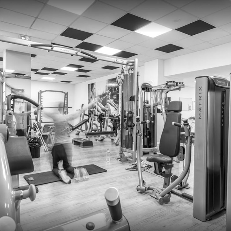 Salle de Sport Matrix : Entraînement et Machines de Musculation Scène intérieure de salle de sport en noir et blanc avec équipement MATRIX, une personne en mouvement flou faisant un exercice au sol, et des affiches murales.